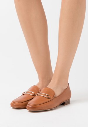 Mocassins en cuir marron avec un embout arrondi, présentant un accent métallique sur le devant et un petit talon carré. Texture lisse et design minimaliste.