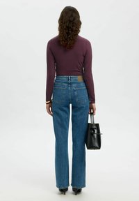 Langere mouwen, fitted bordeaux shirt gecombineerd met hoog-gewassen, blauwe spijkerflared jeans. Een zwarte handtas met knoopdetail wordt in de hand vastgehouden.