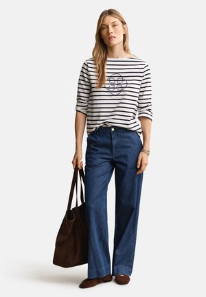 Sieviete, kas valkā baltu un melnu svīrainu garām piedurknēm krekls ar apļveida dizainu, zilas platām kājām bikses, brūnas kurpes un tur lielu brūnu somu.