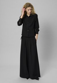 Melna krekla ar garām piedurknēm un redzamām pogām kombinācija ar brīviem melniem plisētiem biksēm, abi izgatavoti no mīksta auduma.