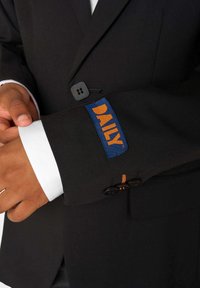 Isik, kes reguleerib musta ülikonna jope mansetti, mille varrukal on oranž "DAILY" silt, näidates valget särgi mansetti all.