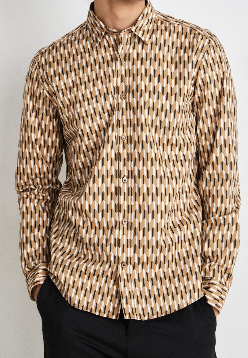Homme portant une chemise à manches longues avec un motif géométrique beige, mains dans les poches d’un pantalon noir, torse et cou visibles.