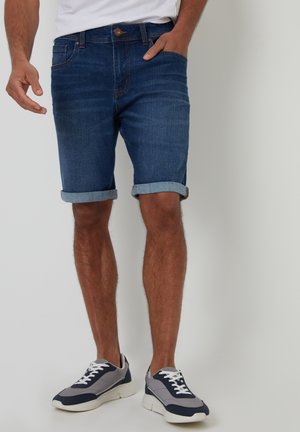 Mørkeblå denim shorts med opkrøllet kant; har en knap- og lynlåslukning, sidelommer og en glat overflade. Bæres med sportssko.