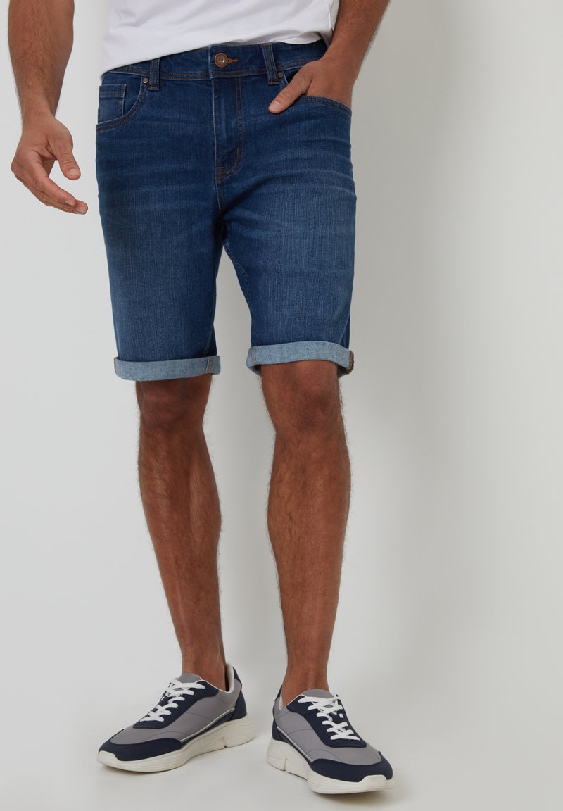 Shorts in denim blu scuro con orlo arrotolato; dotati di chiusura a bottone e cerniera, tasche anteriori e una texture liscia. Abbinati a scarpe sportive.