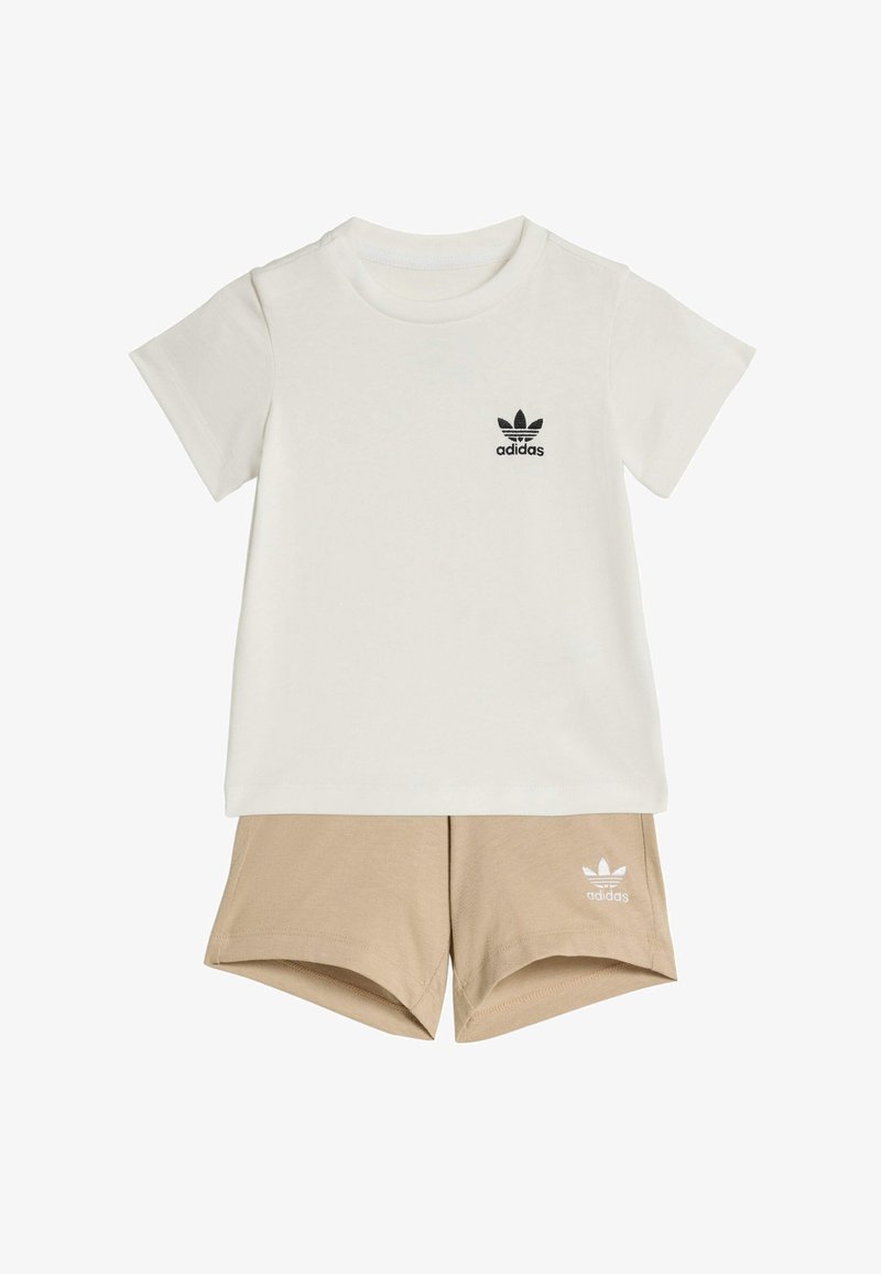 Bijela pamučna majica kratkih rukava s crnim Adidas logotipom i bež kratke hlače s bijelim Adidas logotipom. Obje imaju ležeran kroj.