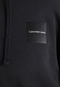 Närbild av en svart huvtröja med en fyrkantig patch som visar texten "Calvin Klein Jeans" på bröstet och en synlig svart snodd.