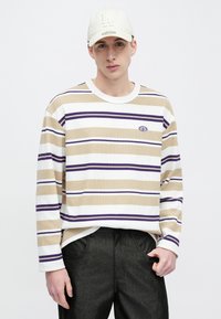 Prugasta majica dugih rukava u bež, bijeloj i ljubičastoj boji, s rebrastom teksturom i logotipom na prsima, uparena s tamnim hlačama.