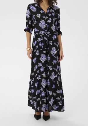 Frau trägt ein schwarzes, langes Blumenkleid mit lila Blumen, vorne geknöpft, Taillengürtel, Dreiviertelärmel und schwarze spitze Absätze.