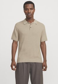Camisa polo de punto beige con mangas cortas, tapeta de tres botones y ribete acanalado. Combinada con pantalones grises holgados.