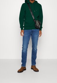 Sudadera verde con capucha, jeans de mezclilla azules, botas de tobillo marrones y un bolso cruzado negro. Atuendo casual con tejido texturizado y diseño sencillo.