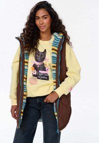 Pruun tikitud vest erksate kudumiste äärtega, mille all on kollane särk, millel on musta kassi ja lilledega graafika.