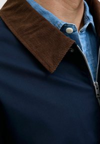 Marinejakke med en brun kordfløjskrave, der har en sølvfarvet lynlås og en lyseblå denimskjorte under.