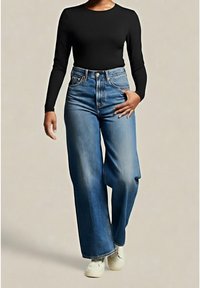 Femme portant un haut noir à manches longues, un jean bleu taille haute à jambes larges et des chaussures blanches, debout devant un fond uni.