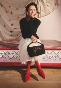 Kvinna som sitter på en dekorerad bänk, klädd i svart tröja, vit lager-på-lager-kjol, röda strumpbyxor och skor, och håller en svart handväska med röd broderi.