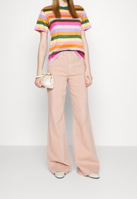 Gestreept T-shirt in paars, groen, oranje en roze; gecombineerd met lichtroze wijde broekspijpen. Model houdt een kleine witte handtas vast.