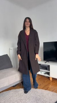 Hosszú fekete kabát sima textúrával, hajtókás gallérral és laza szabással, bordó felsővel és bővülő szárú farmerrel párosítva.