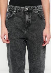 Magas derekú fekete farmernadrág, mely kopott hatású textúrával, öt zsebes kialakítással és a varrások mentén kontrasztos varrással rendelkezik.