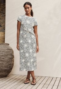 Maxikleid mit einem schwarz-weißen abstrakten Muster, kurzen Ärmeln, einem geknoteten Vorderteil und aus leichtem Stoff gefertigt. Schwarze flache Sandalen.