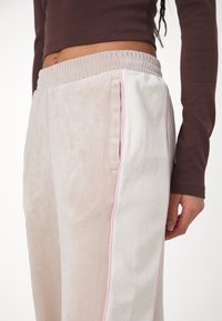 Detailní záběr na osobu, která má na sobě béžové a bílé sametové kalhoty s růžovým lemováním a elastickým pasem, doplněné o hnědý top s dlouhým rukávem.