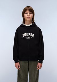 Felpa nera con zip caratterizzata da un logo bianco stampato e l'anno sul petto, abbinata a pantaloni verde oliva. Tessuto morbido, vestibilità rilassata.