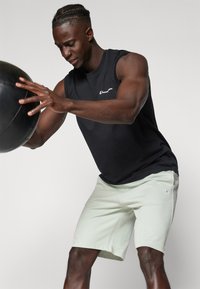 Black sleeveless athletic shirt, light green shorts with logo, textured fabric, individual lifting a black medicine ball against a neutral backdrop.