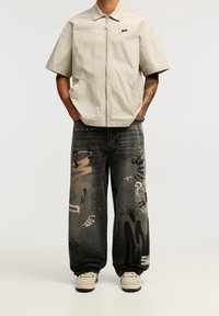 Ljusbeige zip-up jacka med korta ärmar, kombinerad med svarta jeans med guldgraffiti mönster och flera fickor. Lätta skor.