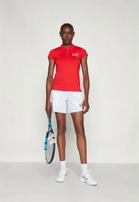 Κόκκινο πουκάμισο polo με γιακά και λογότυπο EA7, συνδυασμένο με λευκά σορτς. Το μοντέλο κρατάει μια ρακέτα τένις, φορώντας λευκά αθλητικά παπούτσια και κάλτσες.