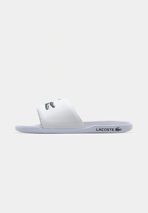 Biele šľapky s textúrovaným vrchom, s výrazným čiernym logom Lacoste a plochou podrážkou. Minimalistický dizajn a ľahký materiál.