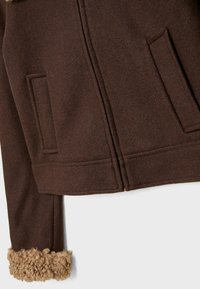 Brun uldjakke med to forlommer og beige krøllet pelskant ved ærmet, vist fra taljen til ærmeslutt.