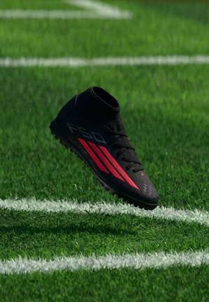 Chaussure de football noire avec des rayures rouges flottant au-dessus d'un gazon artificiel vert près des lignes blanches du terrain.