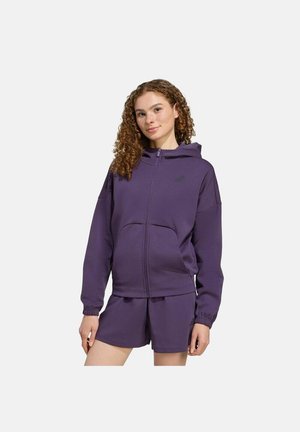 Jonge vrouw met krullend haar, gekleed in een paarse hoodie met rits en bijpassende shorts, staand tegen een effen lichte achtergrond.