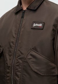 Gros plan d'une veste marron avec une fermeture éclair frontale, des poches rabat et un patch rectangulaire portant l'inscription "Schott NYC" sur le haut de la poitrine.