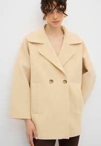 Cappotto oversized beige realizzato in tessuto morbido, presenta un colletto a risvolto, chiusura a doppio bottone e due tasche frontali. Maniche lunghe e ampie.