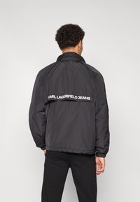 Juodas lengvas švarkas su aukštu apykakle ir elastinėmis rankovėmis. Ant nugaros yra didelis baltas "KARL LAGERFELD JEANS" logotipas.