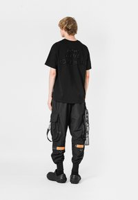 Camiseta negra con "MOVIMIENTO MOD WAVE" en letras texturizadas en la parte posterior. Combinada con pantalones cargo negros y zuecos oscuros de goma.
