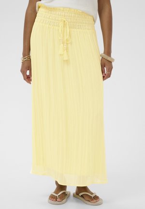 Naine, kes kannab pikkade, helekollaste seelikuga, millel on elastne vöökoht ja tutiga nöör, koos beežide plätude ja käevõrudega mõlemal randmel.