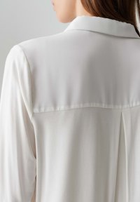 Weiße Bluse mit glatter Textur, die einen klassischen Kragen und ein Schulterblattdetail auf der Rückseite aufweist. Einfaches Design mit langen Ärmeln.