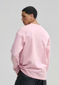 Hosszú ujjú rózsaszín póló, puha anyagból készült, kerek nyakkivágással és laza szabással. Részletei között szerepel egy kis címke a hátsó részen.