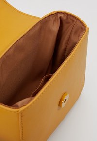 Sac à main en cuir jaune avec un extérieur lisse. Le tissu intérieur est marron. Il dispose d'une fermeture à pression avec un détail de bouton circulaire.