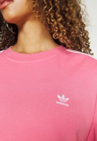 Rosa Adidas sweatshirt med rund hals, med en vit logotyp och ränder på axlarna, tillverkad av strukturerad bomullstyg.