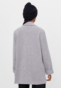 Grauer Hahnentritt-Mantel mit strukturierter Silhouette, Reverskragen und hochgekrempelten Ärmeln, getragen über schwarzen Hosen und einer schwarzen Mütze.