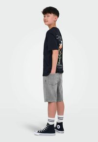 T-shirt noir à manches courtes avec un motif floral et du texte au dos, shorts en denim gris, et baskets montantes noires avec des chaussettes à rayures blanches.