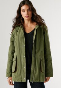 Zaļa āra jaka ar rāvējslēdzēju un tapa aizdari, lielām priekšējām kabatām un kontrastējošu brūnu apkakli. Mīksts, izturīgs audums.