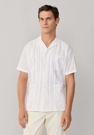 Homme portant une chemise blanche à manches courtes avec des rayures texturées verticales et un pantalon de couleur claire, debout devant un fond uni.