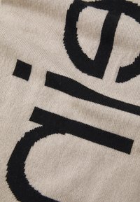 Strikket stoff i kremfarge med fet svart tekst og grafisk design. Glatt tekstur, med distinkte former og mønstre gjennomgående.
