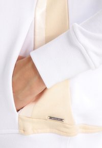 Sudadera blanca con un bolsillo interior beige de suave textura y una pequeña etiqueta de metal que muestra el nombre de la marca.
