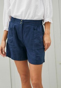Shorts in lino blu navy con cintura elastica, due tasche laterali e due bottoni frontali. Tessuto liscio, design alla lunghezza del ginocchio.