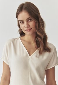 Blusa a maniche corte in tessuto beige chiaro con scollatura a V. Tessuto morbido, vestibilità rilassata e design semplice senza fantasie o embellishments.