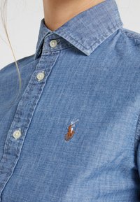 Camisa de mezclilla azul claro con un collar clásico, cierre de botones y un logo bordado en marrón. Textura suave con un sutil patrón tejido.
