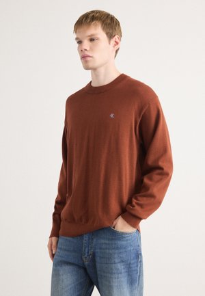 Bruine gebreide sweater met ronde halslijn, geribde boorden en onderkant, met een klein logo op de linkerkant van de borst. Draagt met blauwe denim jeans.
